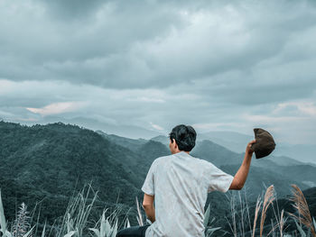 Rear view of man throwing cap against mountain and cloudy sky