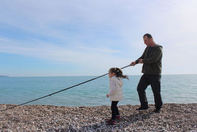 Father and daughter fishing while standing at beach against sky