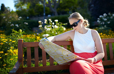 Woman reading map while sitting on bench against plants at park