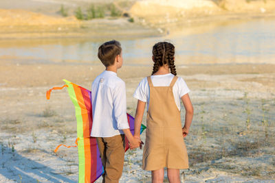 A boy and a girl stand with a kite near the lake and look into the distance.