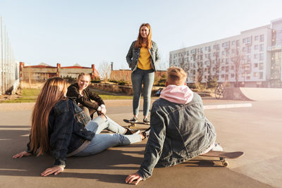 Friends sitting at skateboard park while talking to teenage girl standing against buildings