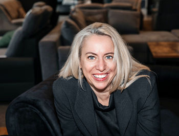 Confident mature woman with platinum hair, smiling in chic black attire surrounded by furniture