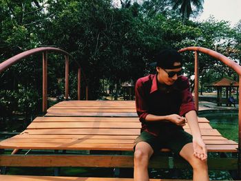 Young man adjusting sleeve while sitting on footbridge over pond at park