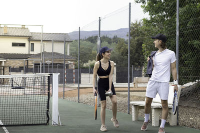 Man and woman looking to each other arriving to a tennis court with their racquets while laughing 