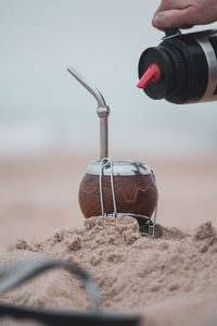 Cropped hand pouring drink in container on sand at beach