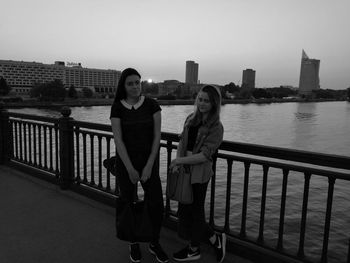 Portrait of young woman by railing against river in city