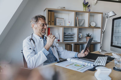 Mature doctor talking on video call over digital tablet while sitting at doctor's office