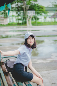 Portrait of smiling young woman sitting on bench in park during rainy season