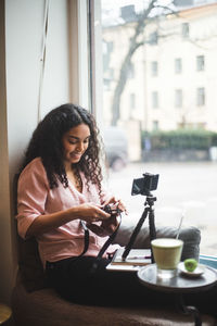 Smiling young female influencer holding camera by window at creative office