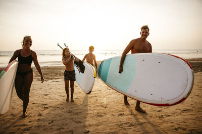 Smiling family with paddleboards walking on sand at beach during sunset