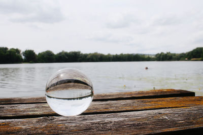 Close-up of glass on table by lake against sky