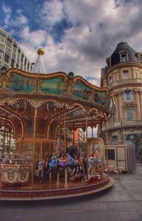 Illuminated carousel by buildings against cloudy sky in city