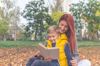 Mother with son reading book while sitting at park during autumn