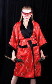 Portrait of young female model holding samurai swords while standing against black curtain