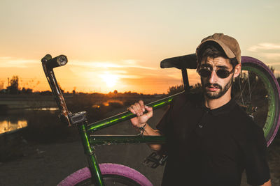 Portrait of young man holding bicycle while standing against sky during sunset