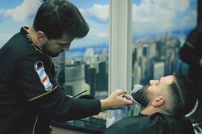 Side view of barber cutting young man beard at shop