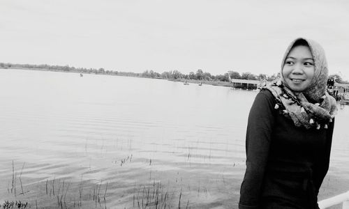 Portrait of smiling young woman standing by lake against sky