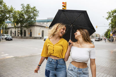 Happy woman holding umbrella with friend walking on street