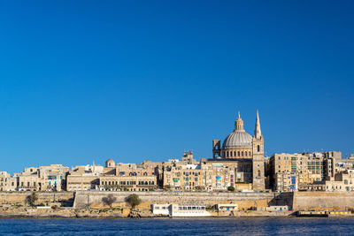 Buildings by river against clear blue sky in city during sunny day