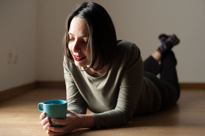 Smiling woman holding coffee cup while lying on floor at home