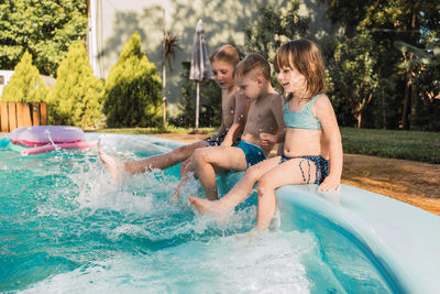 Full body of adorable playful little siblings in swimwear smiling and splashing water of swimming pool with feet while spending time together in backyard of resort on sunny summer day