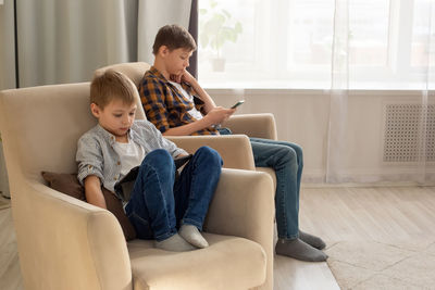 Two boys, sit in beige armchairs, in a room, during the day, play their gadgets