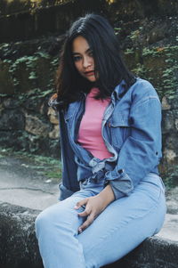 Portrait of young woman wearing denim jacket while sitting on retaining wall