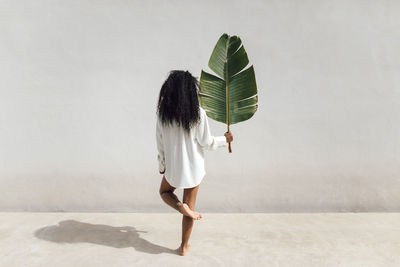Young woman holding banana leaf while standing on one leg in front of white wall during sunny day