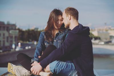 Young couple sitting on retaining wall against sky