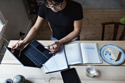 High angle view of young man having sandwich while studying on table at home