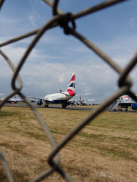 Airplane on runway against sky seen through chainlink fence