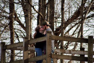 Side view of couple kissing by wooden railing against tree