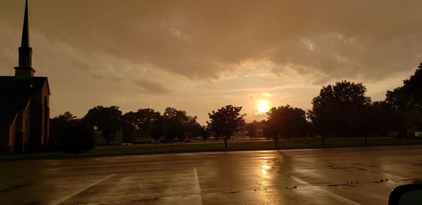 Wet street by trees against sky during sunset