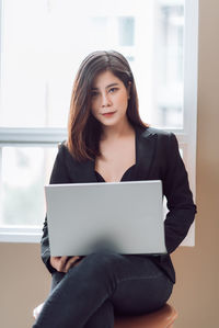 Young woman using mobile phone while sitting on window