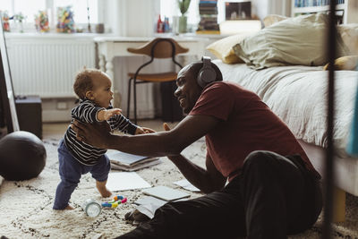 Father holding male toddler while sitting in bedroom at home