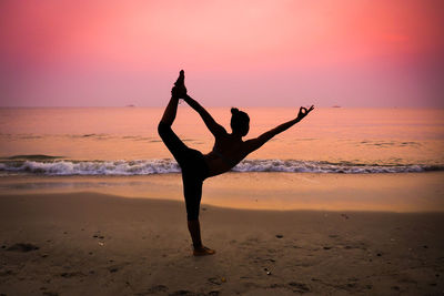 Full length of silhouette woman practicing yoga at beach against dramatic sky during sunset