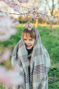 Portrait of a funny little boy wrapped in a blanket enjoying cherry blossoms in a city park.