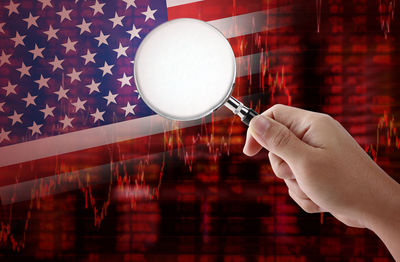 Digital composite image of cropped hand holding magnifying glass against american flag