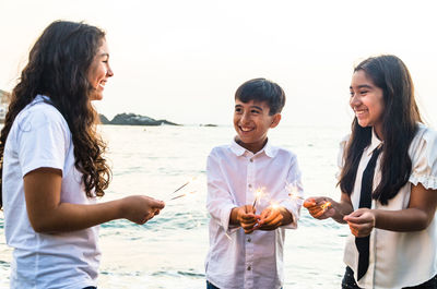 Friends burning sparklers by sea against clear sky
