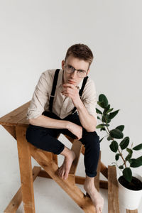 Portrait of young man sitting by potted plant on ladder against white background