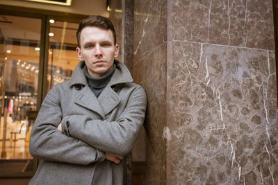 A young man stands outside a shop on the street in a gray coat leaning.