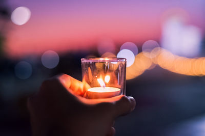 Cropped hand holding lit candle in drinking glass against sky during sunset