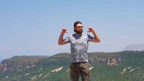 Young man gesturing thumbs up while standing on mountain against clear sky during sunny day