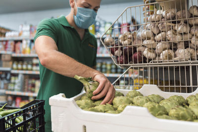 A shopkeeper with a mask taking artichokes from a box in the fruit shop.