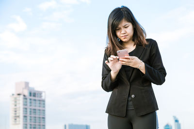 Young businesswoman using mobile phone while standing against sky