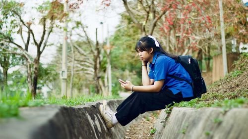 Young woman with backpack using mobile phone while sitting against plants in park