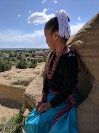 Young woman sitting on rock formation against sky