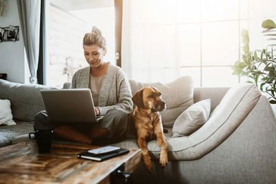 Woman using laptop while sitting with dog on sofa at home