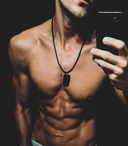 Midsection of shirtless young man taking selfie with smart phone while standing in darkroom