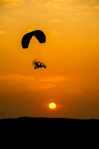 Silhouette person paragliding against orange sky